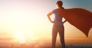 Woman in superhero cape stands facing sunset