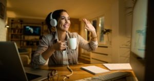 Student with headphones smiles and waves to computer screen while drinking coffee