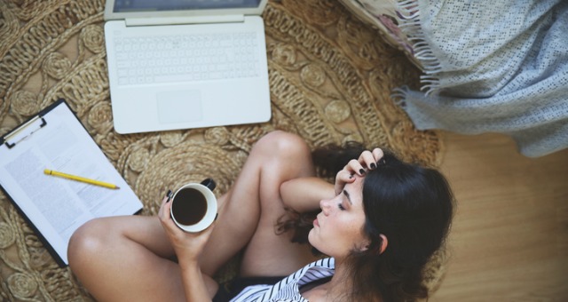 Looking down on girl sitting on floor as she holds coffee cup and puts head in hands in front of computer