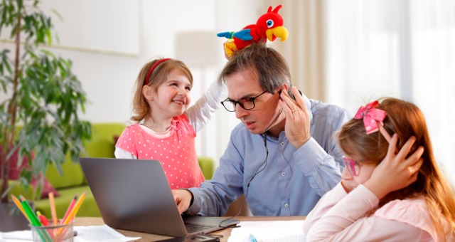 Father of two tries to work at computer while little girls place stuffed animals on his head and other one tries to do homework