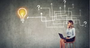 Woman sits on chair with computer laptop with chalkboard behind her of arrows, question marks and a light bulb