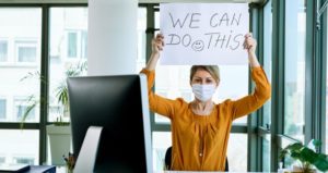Woman has mask on at computer desk while holding up sign that says, "We can do this."