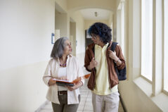 Front view of curly haired man with backpack side by side with mature female teacher and conversing as they approach in hallway.