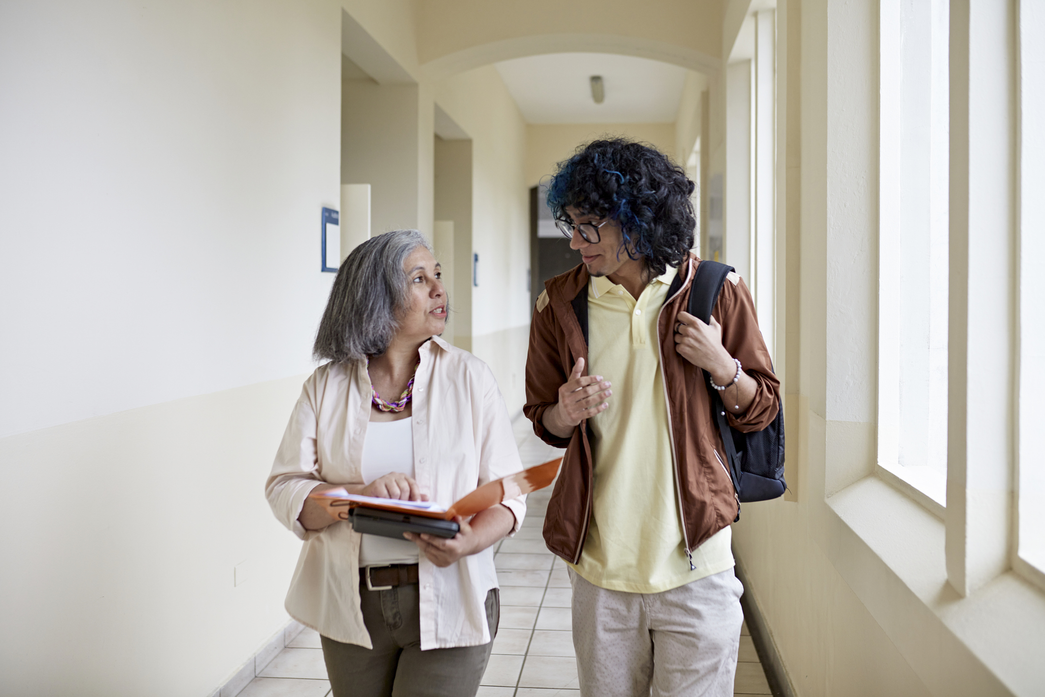 Front view of curly haired man with backpack side by side with mature female teacher and conversing as they approach in hallway.