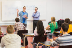 Rear view of students attentively listening to boy by teacher in the classroom