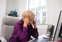 A close-up perspective of a female senior doctor sitting at her desk at work in her examination room in a hospital. She is suffering from work-related stress and is taking a moment to herself as she holds her head in her hands on the desk.