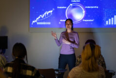 A medium-wide shot of a student standing in front of a projector screen, she is wearing casual clothing. The woman is doing a presentation on data mining at a university campus in London.