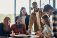 A small group of University students huddle together in class as they plan out and discuss their assignment. They are each dressed casually as they share their ideas with the group.