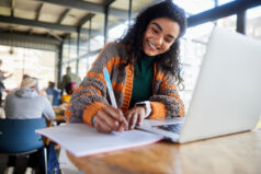 Low angle view of a smiling young female college student writing notes and working on a laptop while doing her assignment in a busy campus cafeteria
