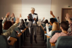 Happy senior female professor choosing one of her students to answer the question on a class at lecture hall. Rapport with students, student participation.