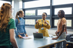 A wide shot of a teacher talking with her students in class, they are wearing casual clothing and discussing what they learned in class, in an authentic way.
