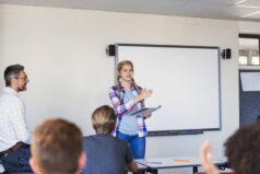 Confident female student standing in front of students and professor. Friends and teacher are looking at teenage girl giving presentation. They are in college classroom.
