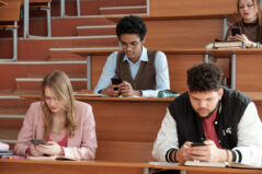Group of contemporary young intercultural students in casualwear sitting by desks in lecture hall and scrolling in smartphones at break