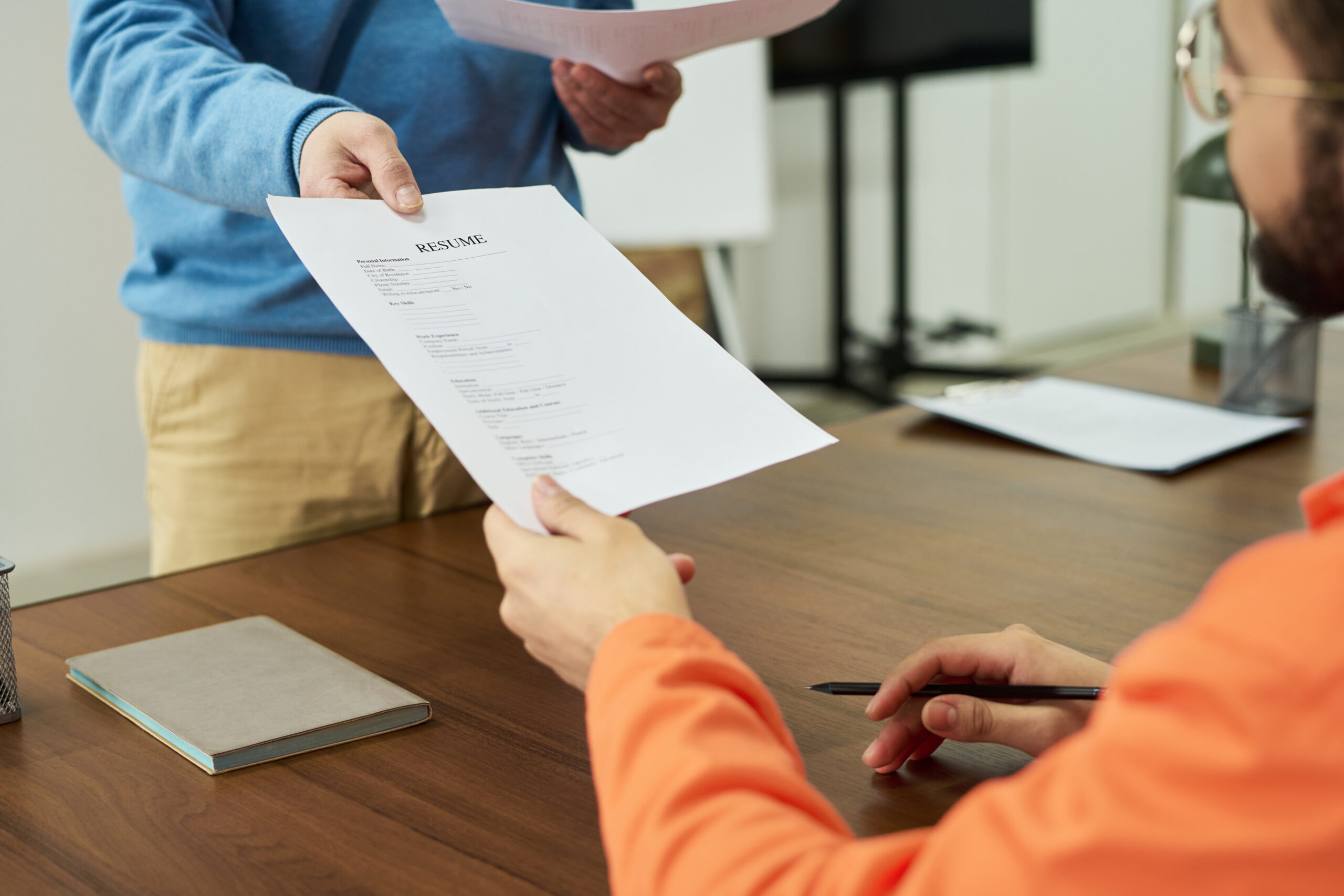 Young man sitting at desk receiving printed resume from standing middle aged man during educational session in prison classroom, both engaging in paperwork exchange
