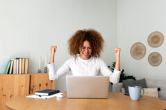 Happy young multiracial Hispanic female celebrating success with arms raised up in front of laptop at home office. Online teaching and learning. Achievement concept.