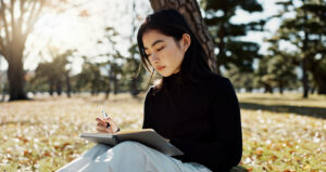 Japanese college student, reading and book in park, thinking and relax by tree, grass or sunshine with peace. Girl, person or student with story in nature, college or idea with vision, knowledge or campus lawn