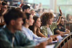 Happy female college student raising her hand to ask a question during a class with her classmates in amphitheater. Filled classroom with happy students.