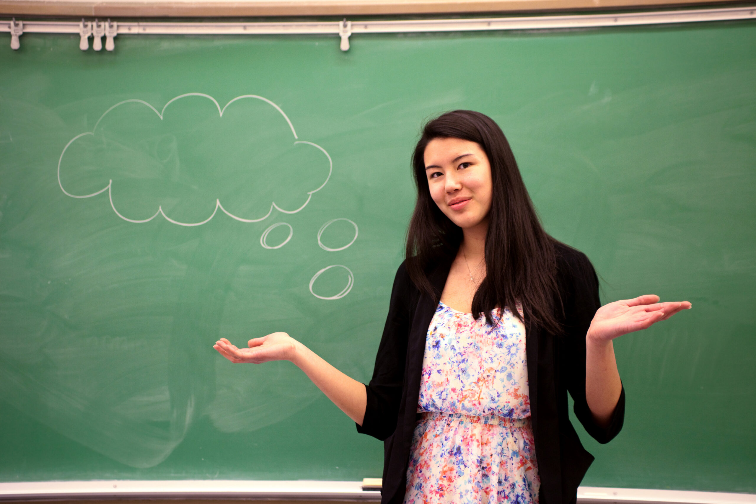 A young woman - a new teacher - stands by a chalk board, wondering with a thought bubble.