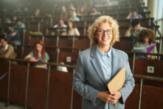 Portrait of happy mature professor standing in lecture hall in front of her students and looking at camera.