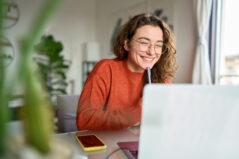 Happy young woman using laptop sitting at desk writing notes while watching webinar, studying online, looking at pc screen learning web classes or having virtual call meeting remote learning from home.