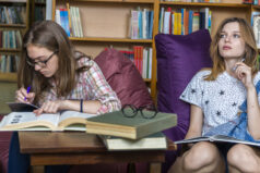 Two teenage girls are writing in the library - one is actively writing while the other ponders.
