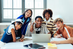 Multiethnic group of young students meeting at the library - diverse teenagers collaborating on project at library, sitting around table with laptop and books, looking at camera. Student wellness. Student wellbeing.