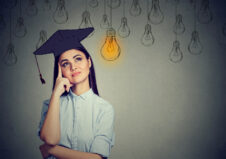 Graduate student young woman in cap gown looking up at bright light bulb thinking isolated gray wall background. Graduation ceremony future career concept