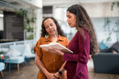 Indigenous educators, one in vibrant attire and the other in professional wear, discuss notes in a modern education office setting with greenery and contemporary furniture, showcasing teamwork and collaboration in a diverse educational environment.